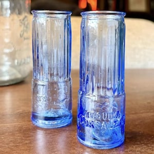 May include: Two vintage blue glass shot glasses with vertical ribbed detailing. The base of one glass reads "TEQUILA CORRALEJO". The glasses are cylindrical and stand on a wooden surface.