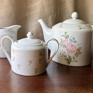 May include: A white porcelain tea set consisting of a teapot with a lid, a lidded sugar bowl with two handles, and a creamer pitcher. Each piece features a delicate pattern of small pink rosebuds and green leaves, with embossed scrollwork.