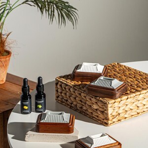 May include: A collection of decorative wooden boxes with white ceramic lids, displayed on a white table and a woven basket. Two small black bottles with the text "Lemon" and "Bergamot" are also present, suggesting essential oils or fragrances.