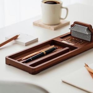 Puede incluir: Un organizador de escritorio de madera con un portalápices incorporado, un pequeño acento arquitectónico y un compartimento para objetos pequeños. Un lápiz, dos bolígrafos y un bloc de notas también están sobre la superficie blanca. Una taza de café está en el fondo.