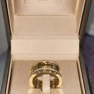 Puede incluir: Un anillo dorado con múltiples bandas, algunas incrustadas con pequeñas piedras brillantes. El anillo se exhibe dentro de una caja de marca con el nombre "BVLGARI" visible en la tapa. El anillo tiene las letras "BVLGARI" grabadas.
