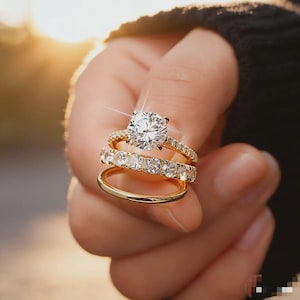 Puede incluir: Un conjunto de tres anillos de oro. El anillo superior presenta una gema grande, redonda y transparente. El anillo del medio está adornado con múltiples gemas claras más pequeñas. El anillo inferior es una sencilla banda de oro.