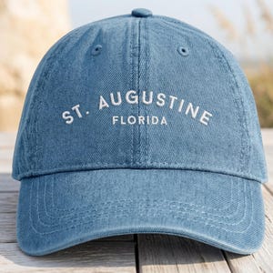 May include: A blue denim baseball cap with the text "ST. AUGUSTINE FLORIDA" embroidered in white. The cap has a curved brim and a button on top. The hat is sitting on a wooden surface.