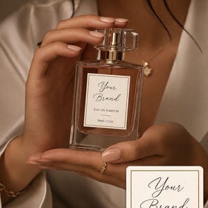 May include: A perfume bottle with a clear glass body and a gold-coloured cap. The label reads "Your Brand, EAU DE PARFUM, 50ml / 1.7oz." The bottle is held in a person's hands. A second label is visible below the bottle.