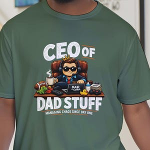Op de afbeelding: Groen t-shirt met de tekst "CEO of Dad Stuff" en een cartoonafbeelding van een man in een stoel. De afbeelding bevat een koffiekopje, een voetbal en een laptop. De tekst "Managing Chaos Since Day One" is ook aanwezig.