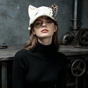 Pode incluir: Boné de beisebol creme com orelhas de gato e estampa de leopardo. O boné tem detalhes metálicos e é usado por uma pessoa com uma camisola de gola alta preta. Fundo escuro, ambiente industrial.