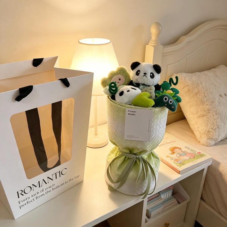 Puede incluir: Una bolsa de regalo blanca con la palabra "ROMANTIC" impresa y un ramo de peluches. El ramo incluye pandas, una flor verde y una rana verde. El ramo est&aacute; envuelto en tela verde.