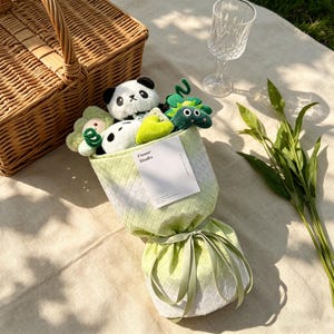Puede incluir: Un ramo de peluches, que incluye un panda, un dinosaurio verde y una flor, envuelto en una tela tejida verde claro y blanca. El ramo est&aacute; atado con una cinta a juego. Una cesta de picnic y una copa de vino est&aacute;n en el fondo.