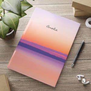 Op de afbeelding: Een notitieboek met een zonsondergangontwerp in oranje, paarse en roze tinten. De naam "Amelia" staat op de kaft. Een zwarte pen en witte oortelefoons liggen op een houten oppervlak.