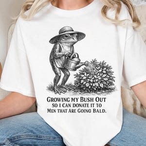 May include: A white t-shirt featuring a black and white line art illustration of a frog wearing a hat and watering a bush. Below the illustration, the text reads: GROWING MY BUSH OUT SO I CAN DONATE IT TO MEN THAT ARE GOING BALD. The shirt has short sleeves.