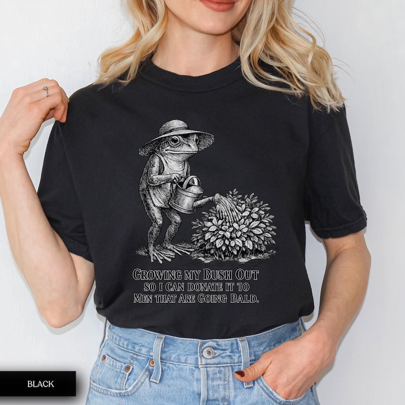 May include: A black t-shirt featuring a white graphic illustration of a frog wearing a sun hat and using a watering can to water a bush. Below the illustration, the text reads: GROWING MY BUSH OUT SO I CAN DONATE IT TO MEN THAT ARE GOING BALD.