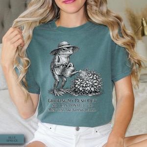 May include: A blue-green t-shirt featuring a black and white graphic of a frog wearing a hat and holding a watering can over a bush. The text below the graphic reads: Growing my bush out so I can donate it to men that are going bald.