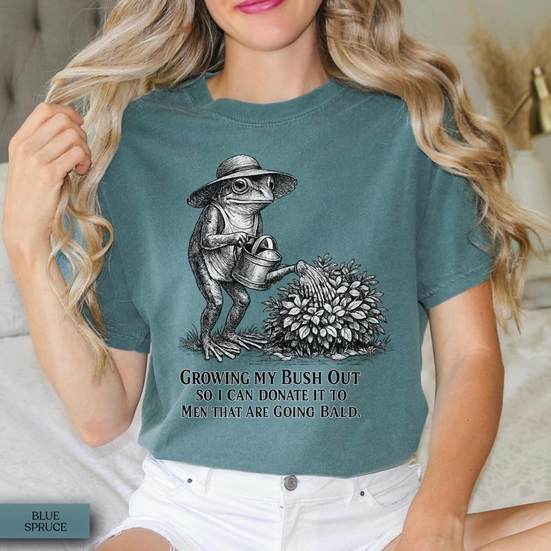 May include: A blue-green t-shirt featuring a black and white graphic of a frog wearing a hat and holding a watering can over a bush. The text below the graphic reads: Growing my bush out so I can donate it to men that are going bald.