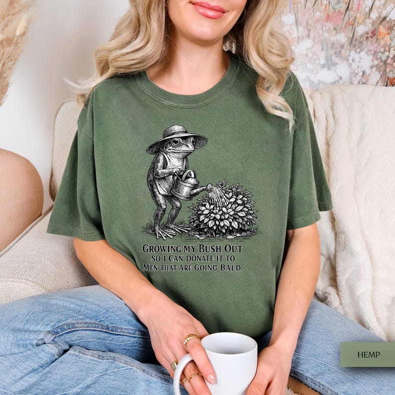 May include: An olive green t-shirt featuring a black and white graphic of a frog wearing a sun hat and watering a bush with a small watering can. Below the graphic, the text reads: GROWING MY BUSH OUT SO I CAN DONATE IT TO MEN THAT ARE GOING BALD.