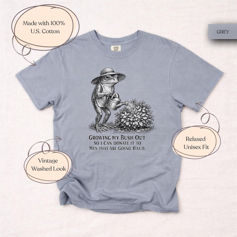 May include: A grey short-sleeve t-shirt with a vintage washed look, featuring a black and white graphic of a frog wearing a hat and watering a bush. The text reads: GROWING MY BUSH OUT SO I CAN DONATE IT TO MEN THAT ARE GOING BALD. Made with 100% U.S. Cotton.