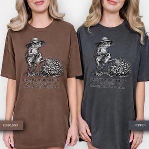 May include: Two short-sleeve t-shirts in espresso brown and pepper gray. Each features a graphic of a frog wearing a hat and watering a bush with a watering can. The text reads: Growing my bush out so I can donate it to men that are going bald.