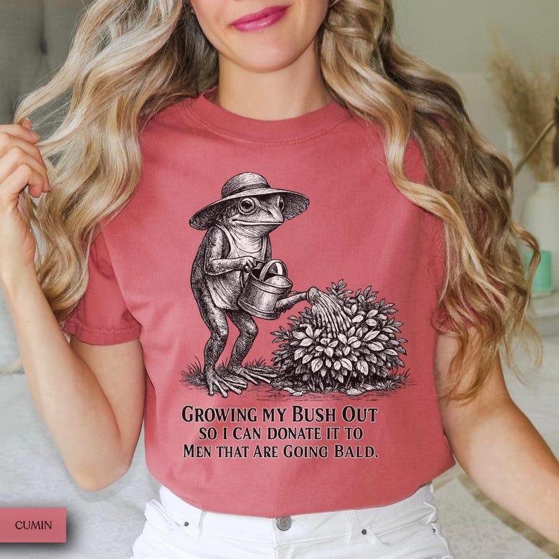 May include: A dusty rose t-shirt featuring a black and white line art illustration of a frog wearing a sun hat and using a watering can on a bush. The text below the graphic reads: Growing my bush out so I can donate it to men that are going bald.