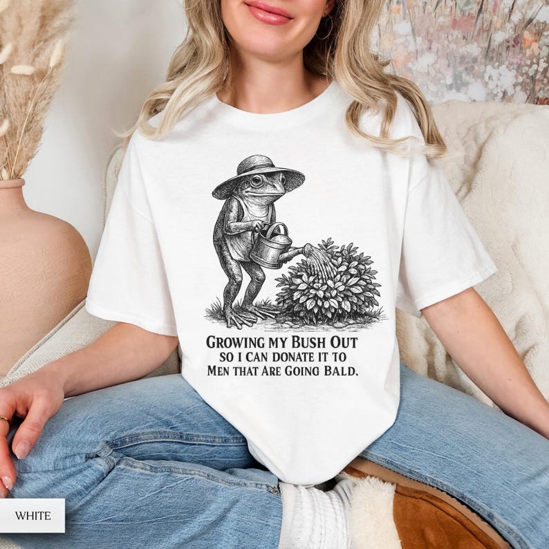 May include: A white t-shirt featuring a black and white line art illustration of a frog wearing a hat and watering a bush. Below the illustration, the text reads: GROWING MY BUSH OUT SO I CAN DONATE IT TO MEN THAT ARE GOING BALD. The shirt has short sleeves.
