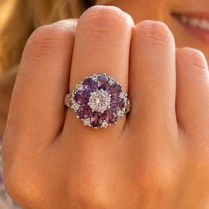 Peut inclure: Une bague argentée ornée d'une pierre centrale ronde et transparente, entourée d'une couronne de pierres violettes taille marquise, avec de petites pierres transparentes sur l'anneau et entre les pétales violets formant un motif floral.