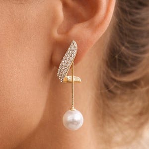 Peut inclure: Boucle d'oreille dorée élégante avec une partie supérieure incurvée ornée de cristaux et une perle blanche pendante. La boucle d'oreille est conçue avec une structure linéaire délicate, créant une esthétique sophistiquée et intemporelle. Un bijou classique.