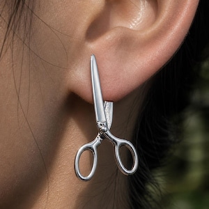 Peut inclure: Boucle d'oreille argentée en forme de ciseaux. La boucle d'oreille est portée sur l'oreille, les lames des ciseaux pointant vers le haut. Le design est détaillé, montrant les poignées et le point de pivot des ciseaux. Un bijou unique.