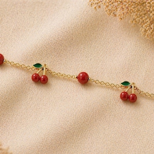 Peut inclure: Un collier doré délicat orné d'un motif répété de breloques cerises rouges et de perles rouges. Chaque breloque cerise comporte deux cerises rouges et des feuilles vertes. Le collier est présenté sur un tissu de couleur neutre.