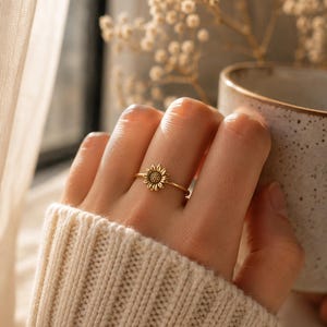Peut inclure: Bague dorée ornée d'un motif de tournesol. La bague est portée au doigt, devant un pull crème. En arrière-plan, une fenêtre et une tasse sont visibles.