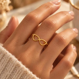 Könnte beinhalten: Ein goldfarbener Ring mit einem filigranen, offenen Rahmendesign in Form einer Katzenaugenbrille. Die Ringschiene ist dünn und poliert und weist eine glatte, metallische Oberfläche auf, die das Licht reflektiert und dezent wirkt.