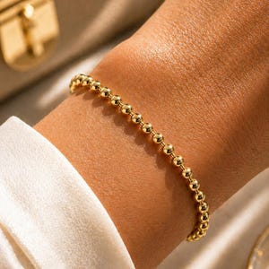 Peut inclure: Un bracelet doré à perles est porté au poignet. Le bracelet est composé de petites perles rondes enfilées ensemble. Le poignet repose sur un tissu blanc. Un sac à main doré est en arrière-plan.