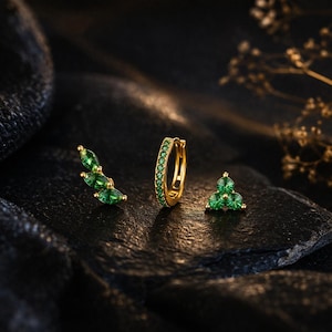 Peut inclure: Trois bijoux dorés ornés de pierres précieuses vertes. L'ensemble comprend un grimpeur d'oreille incurvé, une petite boucle d'oreille et une boucle d'oreille à clous avec un design triangulaire. Les bijoux sont présentés sur une surface sombre et texturée.