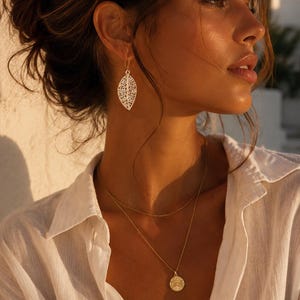 Peut inclure: Une femme porte des bijoux en or, dont une boucle d'oreille en forme de feuille et deux colliers. Les colliers ont une chaîne délicate et un pendentif rond. Elle porte une chemise blanche boutonnée.