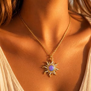 Peut inclure: Collier doré avec un pendentif en forme de soleil. Le pendentif présente une pierre centrale ovale violette, entourée de petits cristaux transparents et d'éléments en forme de rayons de soleil. La chaîne est fine et le collier est porté sur un haut clair.