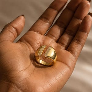 May include: A gold ring with a split design, featuring a band of small, clear stones on each side. The ring is held in an open hand, showcasing its polished surface and intricate details. The ring's design is modern and elegant.