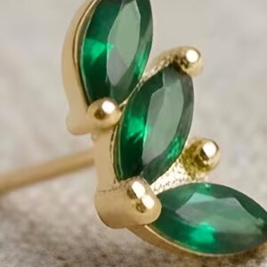 Peut inclure: Boucle d'oreille dorée ornée d'un groupe de pierres précieuses vert émeraude taillées en marquise. Le design comprend de petits accents dorés ronds, créant un arrangement en forme de feuille. L'arrière-plan est neutre.