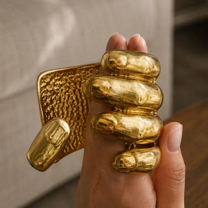 Op de afbeelding: Een goudkleurige manchetarmband met een sculpturaal ontwerp van een hand waarvan de vingers om de pols zijn gewikkeld. Het oppervlak van de armband heeft een getextureerde, gehamerde afwerking en het handelement toont gedetailleerde knokkels en nagels.
