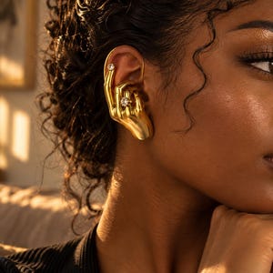Peut inclure: Une boucle d'oreille en forme de main humaine dorée qui s'enroule autour de l'oreille. La main présente une petite pierre transparente sur l'index et une pierre plus grande sur l'annulaire, imitant une bague. La finition est lisse et métallique.
