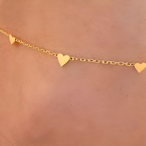 Peut inclure: Un délicat collier doré orné de trois pendentifs en forme de cœur. La chaîne fine soutient les cœurs espacés. Le collier est présenté sur un fond couleur peau, mettant en valeur le design du bijou.