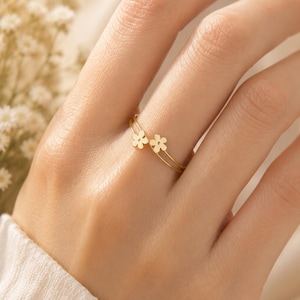 Peut inclure: Une délicate bague en or ornée de deux petites fleurs. La bague a un design à double bande, avec les fleurs positionnées sur le dessus du doigt. La bague est présentée sur une main, avec un fond doux et neutre.