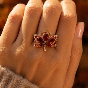 May include: A gold ring featuring a maple leaf design adorned with deep red pear-shaped stones and smaller orange and yellow accent crystals. The leaf motif is centred on the finger, showcasing a vibrant gradient of warm autumn-inspired colours.