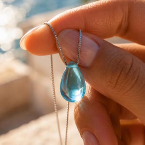 Può includere: Un ciondolo traslucido azzurro a forma di goccia, sospeso a una sottile catenina color argento. Il ciondolo è realizzato in materiale liscio e trasparente che cattura la luce, creando un riflesso luminoso sulla sua superficie arrotondata.