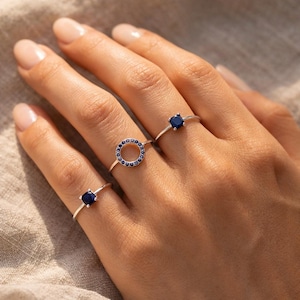 Peut inclure: Trois bagues en argent ornées de pierres bleues. L'une présente une pierre carrée, une autre un cercle de petites pierres, et la troisième une seule pierre carrée. Les bagues sont présentées sur une main avec du vernis à ongles neutre.