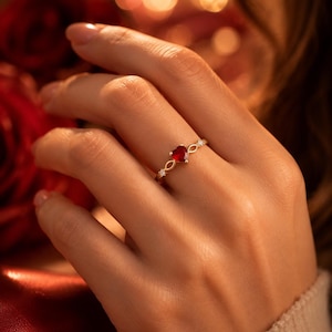 Op de afbeelding: Een gouden ring met een hartvormige, dieprode edelsteen. De ring heeft een delicaat ontwerp met kleine, heldere accentstenen en een oneindigheidssymbool-achtig detail op de band. De ring wordt getoond op een hand, met een vage rode en gouden achtergrond.