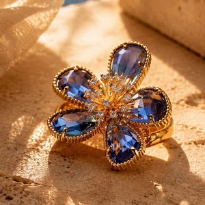 Peut inclure: Une bague dorée ornée d'un motif floral, avec cinq pétales chacun orné d'une grande pierre bleue en forme de goutte. Le centre de la fleur est orné de petits diamants transparents. La bague est posée sur une surface texturée brun clair.