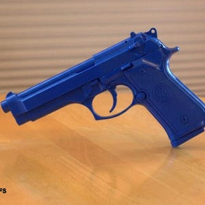 May include: A blue handgun with a textured grip and slide, resting on a wooden surface. The gun features a trigger, hammer, and sights. The text "92FS" is visible at the bottom left.