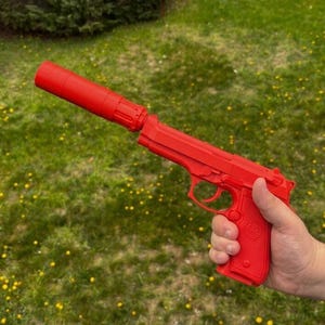 以下が含まれることがあります： 緑の草を背景に、明るい赤色の拳銃とサイレンサーが取り付けられています。 銃器は鮮やかな赤色で、サイレンサーは銃身から伸びています。 銃は人の手で持たれています。