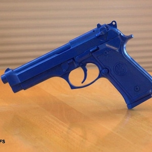 Op de afbeelding: Een effen blauwe replica van een 92FS semi-automatisch pistool. Het model is voorzien van een getextureerde grip met een cirkelvormig embleem, een trekkerbeugel, een slede en een loop. De tekst 92FS staat in het zwart linksonder in de afbeelding.
