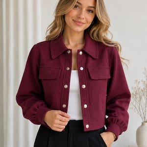 Könnte beinhalten: Burgunderfarbene, kurze Jacke mit Kragen und silbernen Knöpfen. Die Jacke hat zwei Vordertaschen und lange Ärmel. Das Model trägt ein weißes Oberteil und eine schwarze Hose.