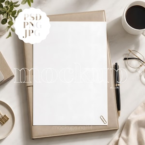 May include: A blank white rectangular paper sheet centered on a beige leather-like portfolio. A black pen, gold paperclip, and gold-rimmed glasses are present. The text PSD PNG JPG appears in a white scalloped circle, and mockup is written across.