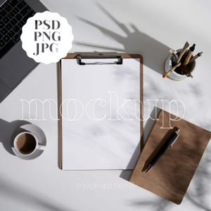 May include: A workspace mockup with a laptop, clipboard, coffee cup, pen, and notebook. The clipboard holds a blank white paper. The image includes the text "PSD PNG JPG" and "mockup" and "mockups no 98".