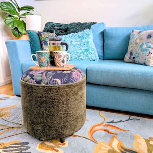 May include: A cylindrical ottoman with a textured olive green fabric base and a dark floral patterned top. A wooden tray sits on the ottoman, holding a glass French press and two ceramic mugs, one with a tropical leaf print and one with a floral design.